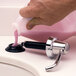 A hand using a Bobrick deck mounted liquid soap dispenser to pour pink liquid into a sink.