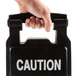 A hand holding a Rubbermaid Executive Series black multi-lingual caution sign.