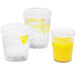 Three Dinex clear plastic tumblers filled with orange juice and garnished with lemon slices on a table with a bowl of lemons.