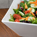 An American Metalcraft square porcelain bowl filled with salad and vegetables on a wood table.