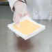 A hand in a white glove holding a piece of cheese on white Patty Paper.