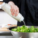 A person using a FIFO Innovations squeeze bottle to pour white dressing onto a bowl of salad.