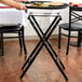 A person using a Lancaster Table & Seating black turned leg tray stand to hold a tray with food on a table with a white tablecloth.