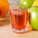 A clear square plastic shot glass filled with liquid next to apples.