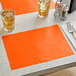 A bittersweet orange scalloped paper placemat on a table with glasses of liquid and silverware.