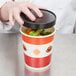 A hand opening a Choice paper soup container filled with vegetables.