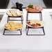 A Tablecraft BKR4 square riser set on a table with a plate of cheese, grapes, and bread.