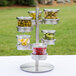 A Cal-Mil stainless steel rotating display stand with three jars of food with hinged lids.