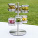 A Cal-Mil stainless steel tiered stand with jars of food on a table.