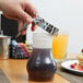 A hand using a Tablecraft syrup dispenser to pour syrup into a Tablecraft jar on a table.
