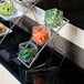 An American Metalcraft chrome three-tier display stand holding glass containers of broccoli and baby carrots on a counter.