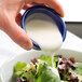 A person pouring navy blue Thunder Group fluted ramekin of white sauce onto a salad.