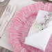 A pink Charge It by Jay glass charger plate on a table with a napkin and silverware.