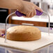 A hand uses the purple-handled Wilton Small Cake Leveler to cut a round cake.