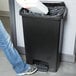 A person using a Rubbermaid Slim Jim black step-on trash can to throw away a box.