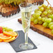 A Fineline Flairware clear plastic champagne flute filled with champagne on a table next to a plate of orange slices.