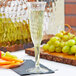 A Fineline clear plastic champagne flute filled with champagne sits on a table next to a plate of fruit.