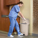 A nurse in blue scrubs and gloves uses a Rubbermaid Slim Jim beige plastic trash can with a step-on lid.