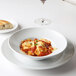 A Tuxton bright white china bowl filled with pasta with sauce on a table with a plate of bread and a glass of wine.