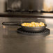 A round black American Metalcraft silicone trivet with a pan of macaroni and cheese on a stove.