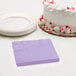 A stack of lavender Creative Converting beverage napkins on a table with a cake on a white plate.