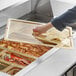 A hand using a Cambro amber plastic lid with a spoon notch to open a tray of food.