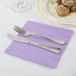 A plate of food and silverware on a purple Creative Converting Luscious Lavender napkin.