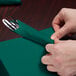 A person's hands wrapping a hunter green Creative Converting luncheon napkin around silverware.