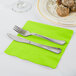 A knife and fork on a Fresh Lime Green napkin on a table next to a plate of food.