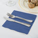 A silver fork and knife on a navy blue napkin next to a plate of food.