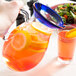 A hand holding a Libbey Aruba pitcher of orange and lemonade over a table with a blue rimmed glass of orange liquid.