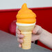 A hand holding an orange ice cream cone with a yellow butterscotch shell coating swirl.
