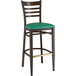 A Lancaster Table & Seating metal ladder back bar stool with dark walnut wood grain finish and green vinyl seat.