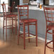 A group of Lancaster Table & Seating Spartan metal bar stools with mahogany wood grain and red vinyl seats next to a bar counter.