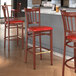 A Lancaster Table & Seating metal bar stool with a mahogany wood grain finish and red vinyl cushion.
