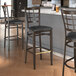 A Lancaster Table & Seating metal bar stool with a dark walnut wood grain finish and black vinyl seat.