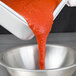 A person pouring red sauce from a Vollrath stainless steel steam table pan into a bowl.