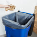 A hand throwing a black plastic bag into a blue Rubbermaid trash can.