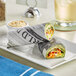 A plate of food wrapped in Choice newspaper deli wrap on a table.