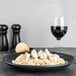 A black Hall China oval platter with pasta, chicken, and herbs on a table with a glass of wine.