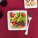 A Libbey Ultra Bright White square porcelain bowl filled with salad with tomatoes and croutons with a fork on a table.