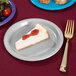 A slice of cheesecake on a Creative Converting shimmering silver paper plate with a fork and strawberry jam.