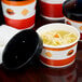 A table with several black and white striped paper soup containers with lids, filled with soup and one with noodles.