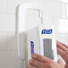 A person using a white GOJO wall plate for an ADX-7 hand sanitizer dispenser on a wall.