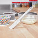 A Tablecraft white spatula in a bowl of frosting with plastic containers of sprinkles.