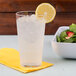 A clear GET plastic tumbler of water with ice and a lemon slice on a yellow napkin.