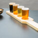 A row of Libbey Straight Sided Tasting Glasses on a natural flight paddle filled with beer.