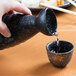 A hand pouring water into a black Whittier Nagoya stoneware sake bottle.