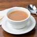 A Tuxton eggshell china bouillon cup filled with soup on a plate with a spoon and crackers.