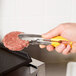A person holding a meat patty with Vollrath stainless steel tongs with a yellow handle.